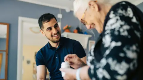 Getty Images A man with dark hair and a short beard sitting talking to a woman with grey hair and glasses. The woman appears to be holding a medicine container. Both are smiling.