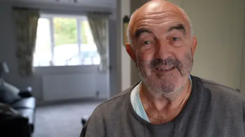 A man with a balding hair line and grey stubble stands in a house, with a lounge and windows behind him to his left. He is smiling and wearing a grey top and a gold chain necklace.