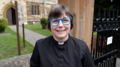 A woman with short dark hair is wearing bright blue-rimmed glasses. She is smiling and dressed in all black, apart from a white dog collar. She stands outside the gates to a church; the arched door of which can be seen open at the end of a path behind her.