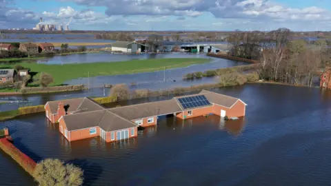 Getty Images A village is almost entirely flooded by blue waters stretching almost to the horizon. In the foreground, a large red-brick bungalow property is surrounded by water. In the middle-distance are more homes and farm buildings, with patches of grass and trees. In the background, a large power station sits on the horizon under a blue sky.