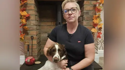 Katy Allinson A woman with short blonde hair and glasses is kneeling in front of a fire place. She is wearing a black T-shirt and black bottoms and is cuddling a white springer dog with brown markings.