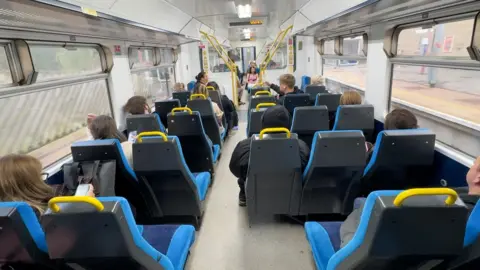 Black and blue train seats face away from the camera down a train carriage. Some of the seats have yellow grab bars. Around half the seats have passngers in them facing away from sight. On either side there are windows, and outside the platforms of a station are visible on both sides of the picture.