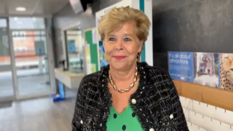 A woman with short brown hair and a green top with large black spots on it and a black coat, stands in front of Goole Rail station's ticket office. She has lipstick on and is smiling. She wears a think gold-coloured necklace, and behind her is the ticket office counter, and the glass doors to the platforms.