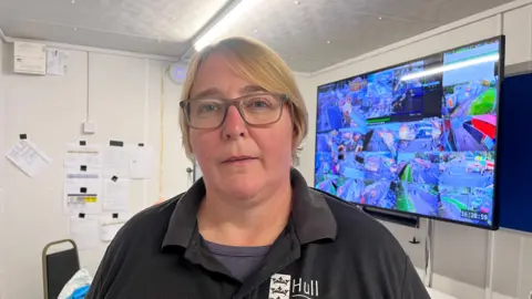 BBC A woman with blond hair and glasses stands in front of a large screen showing footage from a number of CCTV cameras. She is wearing a black t-shirt with the Hull City Council logo on it.
