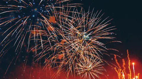 Getty Images Starbursts of orange and yellow fireworks against a dark sky, with red smoke and lights rising to the right.