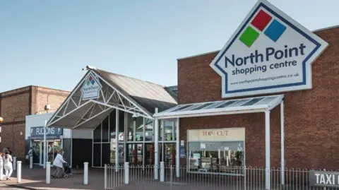 Google The image shows the main entrance of North Point Shopping Centre. Above the entrance is a large sign displaying the name 'North Point' alongside a logo made of three diamonds in red, green, and blue. The building has glass doors and windows. Two shops are visible: 'Fultons' on the left and 'Top Cut' on the right. A person is pushing a trolley near the entrance and there is a silver metal fence running across outside.