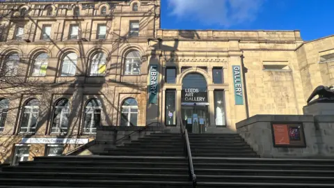 Exterior view of Leeds Art Gallery, featuring a stone façade with arched windows, a wide staircase leading to the entrance, and vertical banners labelled "LIBRARY" and "GALLERY".