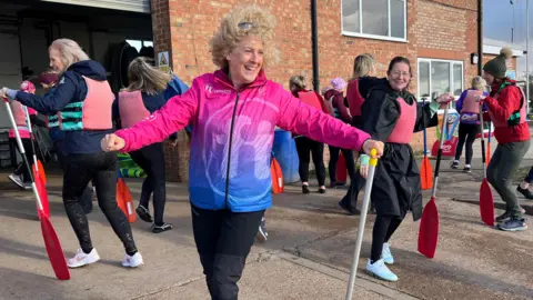 Sam Barlow with wild, curly blonde hair being blown by the fresh breeze is wearing a pink and blue Fitmums and Friends tracksuit top. She's got a canoe paddle in her left hand and circles it as if it's an elegant cane in a Fred Astaire dance as she leads a group of women laughing women in a pre paddle warm up at Welton Waters boathouse.