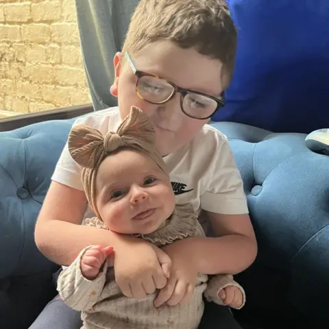 Supplied A five-year-old boy sits on a blue sofa and holds his baby sister, who is sitting in his arms. The boy has short brown hair and wears dark-framed glasses and a white T-shirt. His sister wears a cream-coloured jumper and matching headband with a large bow. She is smiling.