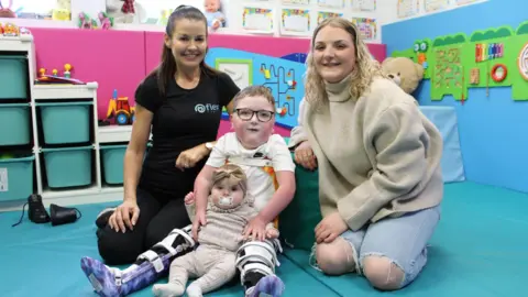 Supplied Two women smile as they kneel on either side of two children - a five-year-old boy and a baby girl - in a children's play area with soft furnishings in blue and pink, toys and storage shelves. To the left is therapist Lisa Speight, a woman with dark-brown hair, tied back, wears a black top with the word "flex" in blue letters, and black trousers. To the right, a woman with long blond curly hair wears a baggy cream jumper and light-blue ripped jeans. 