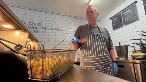 Kevin Shoesmith/BBC A man with short, ginger hair dips a basket containing chips into a fryer in a fish and chip shop.