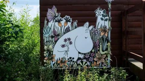 Getty Images A drawing of a hippo-like creature stuck on to the side of a wooden dark brown hut. The creature has a black outline and a white body and is holding four purple flowers in its hand. It's eye is closed. Around it are colourful wildflowers that have been drawn with a butterfly and caterpillar with animated faces.