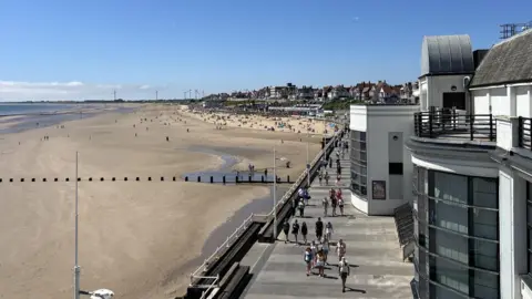 East Riding of Yorkshire Council A high view of a promenade with large modern buildings next to it. The sky is blue and sun is out and several people can be seen walking along it. A beach is located next to it with the tide out and houses next to the beach where several people are dotted about.