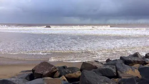 Environment Agency Brown large rocks located on a beach with a sea behind it and waves coming in. The sky is cloudy.