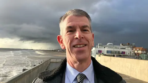 BBC A man with short grey hair is smiling and wearing a black coat, blue shirt and black and white spotty tie. He is standing on a promenade next to a beach. The sky is cloudy.