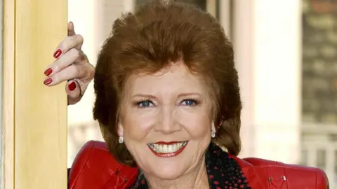 AFP via Getty Images Cilla Black smiles at the camera as she rests her hand on a vertical wooden post. The TV host is wearing a red jacket and a black and red spotted blouse. The red polish on her nails compliments her lipstick. She has short wavy red-hair. 