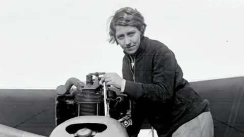 PA Media Black and white photograph of Amy Johnson dressed in flying gear in the cockpit of an aircraft. She is looking towards the camera and smiling, with her goggles pulled up on her forehead.