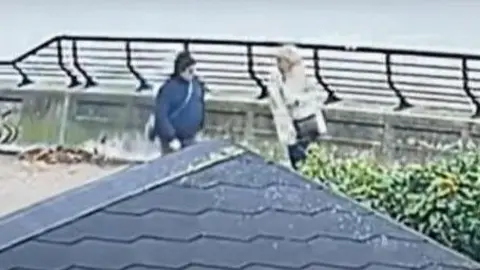 Humberside Police A still image taken from CCTV footage of two people walking along the bank of the Humber estuary. A figure on the right is wearing a dark jacket and what looks to be a black hat. The person on the right of the image has a dark cross body bag, a light coat and light hair.