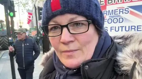 BBC Anna Longthorp stands on the pavement in London with tractors on the road and a banner behind her. She wears glasses and a blue woollen hat and hooded top and a black coat with fur trim.