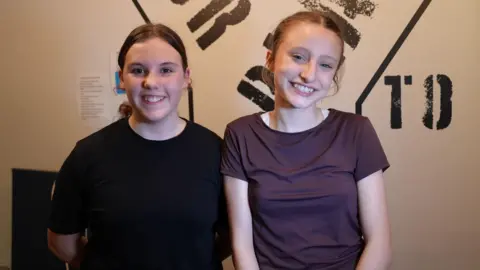 Two young people are pictured standing indoors against a wall featuring bold, stencilled lettering in black on a light background. Both girls are positioned close together, wearing casual dark clothing. Faye on the left is wearing black, and Ayesha has a purple short sleeved top. Both girls have their brown hair tied behind them and are smiling.
