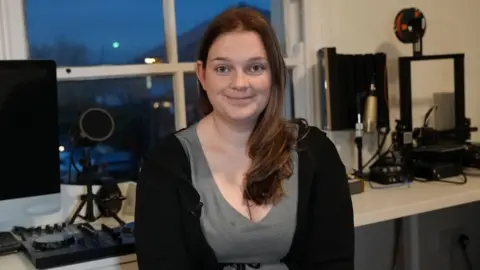 A woman in a grey t-shirt and black cardigan is seated in a well-lit room featuring a desk equipped with various electronic and audio devices. On the left side of the image, a large computer monitor and audio mixer are visible. In the background is a window overlooking Pocklington town centre. Katherine has brown eyes and long brown hair running down her left shoulder and is smiling. 