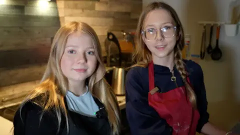 BBC Two young people, Molly and Sinead, are pictured in a modern kitchen setting, standing side by side near a counter top. Both are wearing aprons. Molly on the left has long blonde hair, a light blue T-shirt and a black apron. She is smiling. Sinead has long brown hair, one length in a plait, and is wearing glasses and a red apron. Behind them, there are stainless steel appliances and utensils hanging in a contemporary-looking cooking psace. 