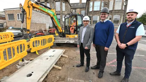 Hull City Council Three men wearing hard hats stand next to a construction site, there are yellow safety barriers and a man in a yellow digger working. 