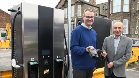 Hull City Council Two men stand together holding a bus charging cable next to a bus charging point. The man on the left is taller, wearing a blue jumper and glasses. The man on the right wears a grey suit jacket and shirt. They are both smiling. 