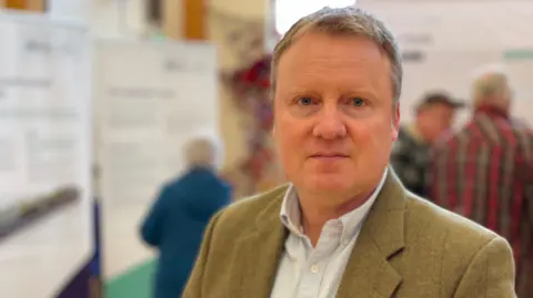 BBC A man with short fair hair looks at the camera. He is wearing a light blue shirt and a brown-green jacket. He is standing in a room full of display boards and people looking at them. He is in focus while the background is out of focus.