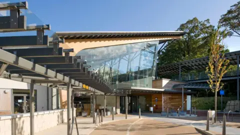 NHS Hull University Teaching Hospitals The entrance to a hospital with a road and path leading up to it. The main building has a slanted ceiling and a large open window either side with two longer buildings either side of it.
