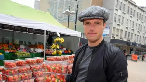 Tommy Coyle Tommy Coyle wearing a flat cap, standing next to the fruit stall in Hull City Centre. He is holding multiple packs of strawberries and is wearing a black zip up jacket.