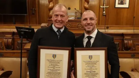 Hull City Council Neil Hudgell and Tommy Coyle receiving their awards at Hull's Guildhall. Both men are wearing dark jackets and a shirt and tie. They are standing in the council chambers holding the awards in front of them.