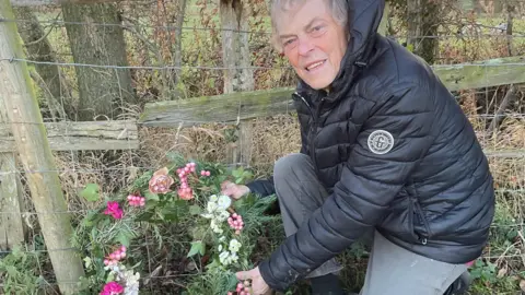 Peter Fall A man in a black puffa jacket and grey joggers lays a floral wreath next to a wooden and wire fence in a grassy field.