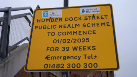 A bright yellow roadside sign with black text is mounted on a metal post near a railing. At the top, there are two logos: one for "Hull City Council" on the left and one for "National Highways" on the right. The sign is outdoors, with a grey overcast sky in the background and part of a metal fence visible on the left. The main text reads: "HUMBER DOCK STREET PUBLIC REALM SCHEME TO COMMENCE 01/02/2025 FOR 39 WEEKS. Emergency Tel: 01482 300 300.”