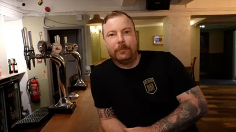 The image shows the interior of a bar with wooden flooring and a polished wooden counter in the foreground. On the counter are several chrome beer taps with branded handles. Behind the counter, there is a fire extinguisher mounted on the wall and shelves holding bottles. The lighting is warm. A man is leaning on the bar counter, wearing a black T-shirt with a logo. His arms are resting on the counter, and both arms are covered in detailed tattoos. In the background, there are cream-colored walls, and a doorway leading to another room. 
