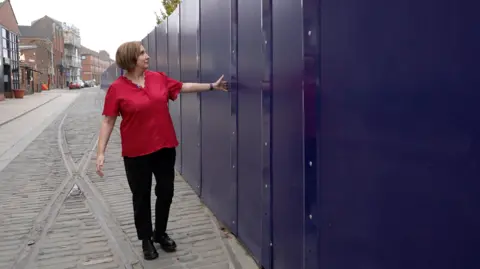 BBC A woman wearing a bright red short-sleeved shirt and black trousers is standing on a cobblestone street next to a tall, solid barrier made of dark blue panels. She is gesturing toward the barrier with one arm extended, as if indicating its length or height. The street has old metal tram lines running along its surface, and older brick buildings are visible in the background on the left side. 