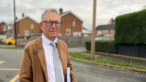 BBC A man with shot grey hair stands in a residential street with two tall telegraph poles in the background. He wears dark-rimmed glasses, a brown jacket, white shirt and silver tie and has a serious expression on his face.