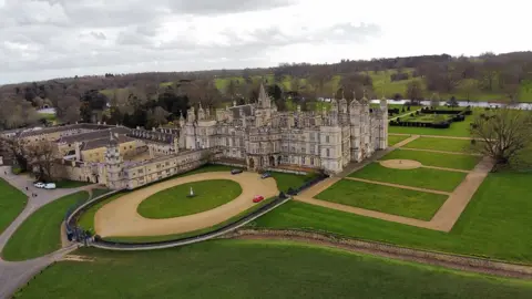 Aerial view of Burghley House in Lincolnshire a three-storey stately home with towers and turrets there are extensive grassed grounds with a large lake and trees in the background