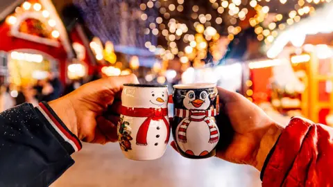 Getty Images Two hands holding festive snowman and penguin mugs with the lights of stalls in the background