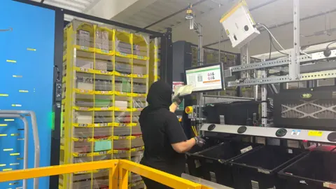 A woman wearing a black headscarf and black T-shirt stands with her back to the camera. She is holding a parcel to a screen. To her left is a large yellow set of shelves. In front of her are black crates and machinery. 