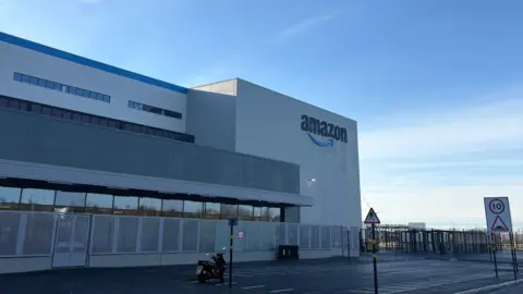 The outside of the Amazon warehouse in North Ferriby. It is a large white building with the Amazon logo on one side and a large car park in front. The sky is blue. 