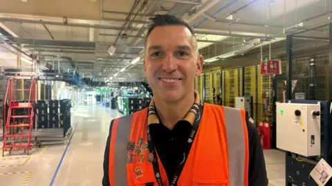 A head and shoulders shot of David Benfell smiling at the camera. He is wearing a black polo shirt with a patterned collar and an orange hi-vis vest with badges on one side. He has short brown hair. He is standing inside a warehouse. 