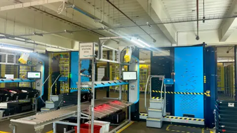 Machinery and large yellow robotic machines behind a cage inside an Amazon warehouse. Various screens can be seen and a sign reads "Universal Station 2314". Black and yellow safety cords are stretched across some areas.