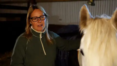 Crispin Rolfe / BBC Sarah Russell has shoulder-length brown hair she wears glasses and a green fleece top. She is standing next to a white horse