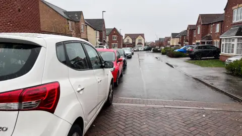 A row of cars can be seen parked all the way down a residential street close to Castle Hill Hospital. There are houses and gardens on either side.