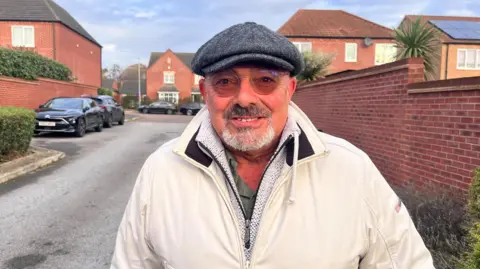 BBC Tony stands in front of some parked cars in a residential street close to Castle Hill Hospital wearing a white coat, flat cap and glasses.