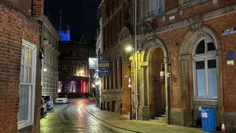 Kevin Shoesmith/BBC A photograph, taken at night, show a cobbled street curving to the right. On the inside bend is a brick-built Victorian-era building. The cobbles are glowing from the reflection of streetlamps.