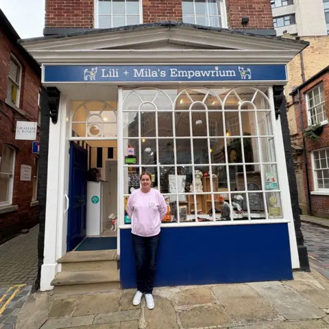 Kevin Shoesmith/BBC A woman in her 20s is standing in front of her pet shop which is painted blue. It has a large glass frontage containing panes.