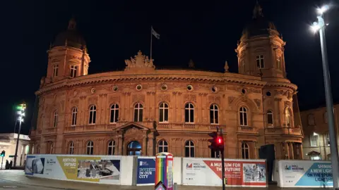 Kevin Shoesmith/BBC A large building, made from sandstone, glows in the light from street lamps. In front of it, are hoardings promoting Hull City Council's significant restoration project.