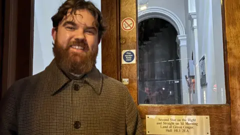 Kevin Shoesmith/BBC A bearded man, in his 20s, is wearing a smart, brown check jacket. He is is smiling next to a door that displays a golden plaque. It says "Second Star on the Right and Straight on 'til Morning, Land of Green Ginger, Hull, HU1 2EA".